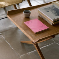 Wooden table with light oak finish, a pink notebook, gray books, and a black cup against a textured stone floor.