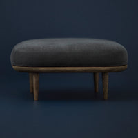 Dark gray upholstered ottoman with natural wood legs against a deep blue background. Smooth finish and modern design.