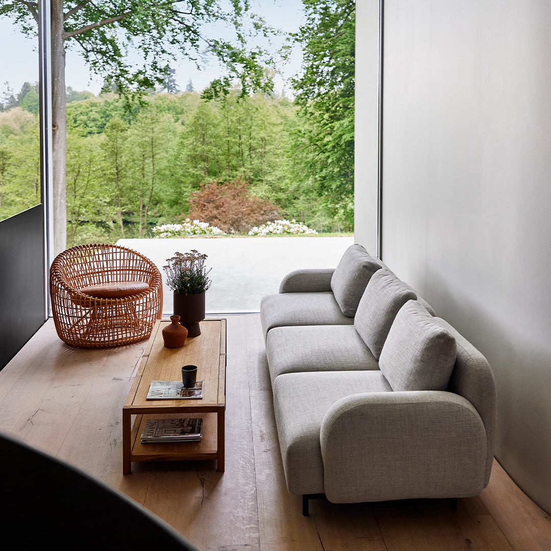 Light gray curved sofa paired with a natural wood coffee table and an earthy-toned rattan chair in a lush green setting.