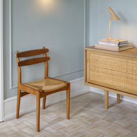 Natural wood chair with a woven seat, complemented by a light blue wall and a textured beige dresser.