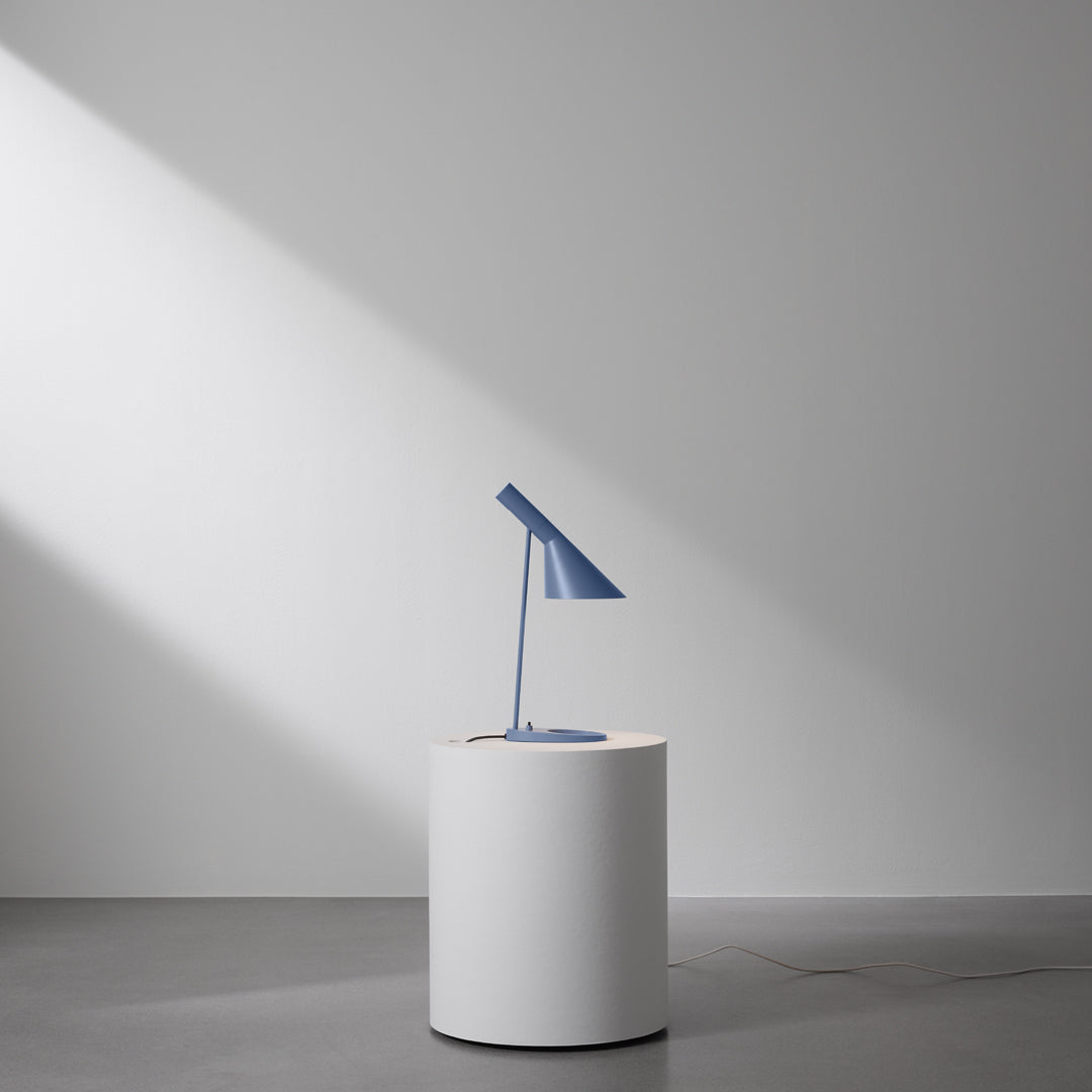 Blue desk lamp on a round white pedestal, set against a minimalist gray background with soft shadows.