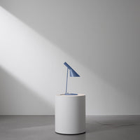 Blue desk lamp on a round white pedestal, set against a minimalist gray background with soft shadows.