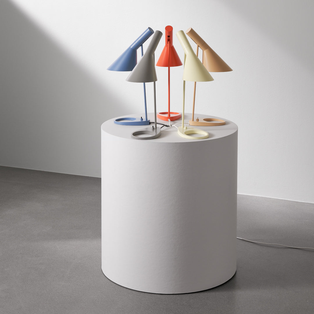 Colorful table lamps in blue, yellow, orange, grey, and white on a round, white pedestal against a minimalist background.