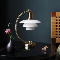 Modern brass table lamp with white layered glass shade against a dark wall, featured on a round black table.