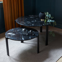 Round black marble coffee tables with white veining, set against a dark green wall and complemented by warm gold curtains.