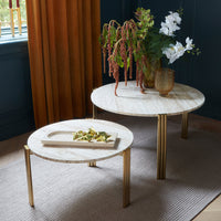 "Two round tables with marble tops and gold legs, accented by greenery and neutral tones in a stylish indoor setting."