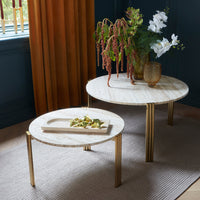 Elegant round marble tables with golden legs, surrounded by greenery and vibrant curtains, on a textured rug.