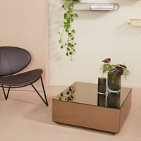"Modern square coffee table with a reflective bronze surface, paired with a sleek black chair and green foliage."