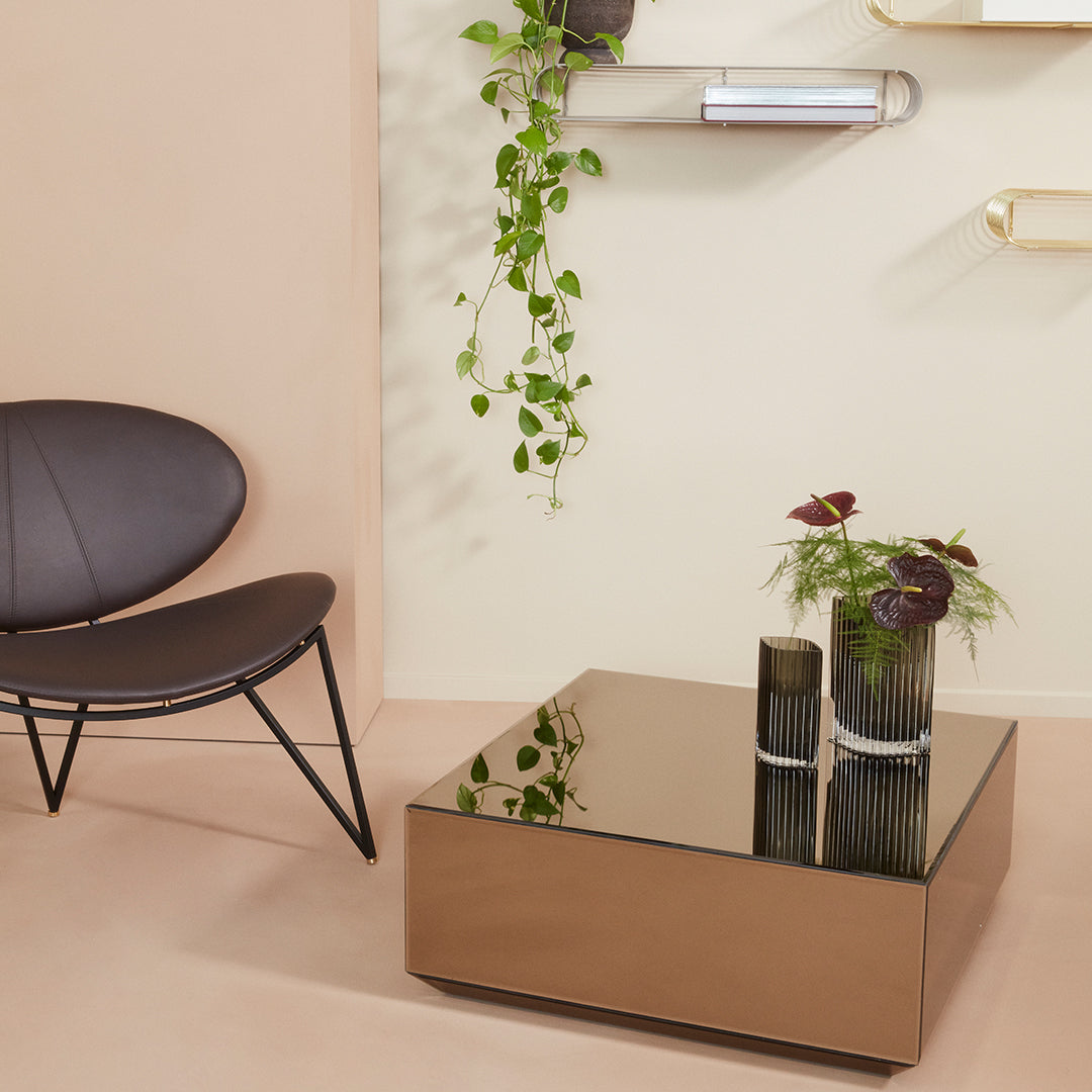 Modern square coffee table with a reflective bronze surface, accented by dark green plant and neutral backgrounds.