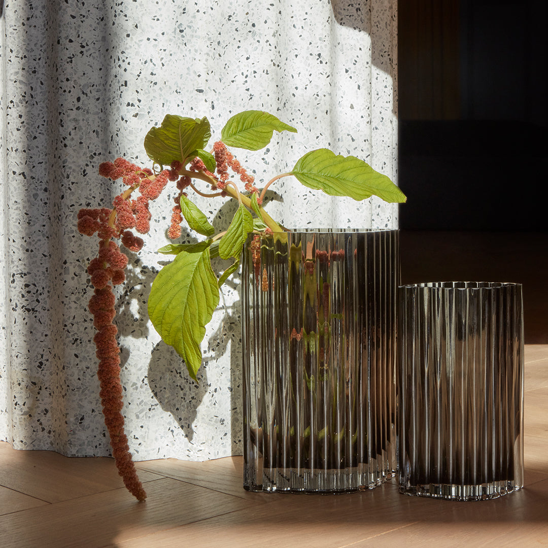 "Clear glass vases with ribbed patterns, featuring green leaves and pink accents against a speckled white backdrop."