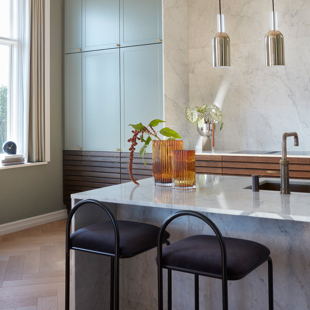 "Modern kitchen with marble countertop, black bar stools, green accents, and amber glass vases, featuring sleek lines."
