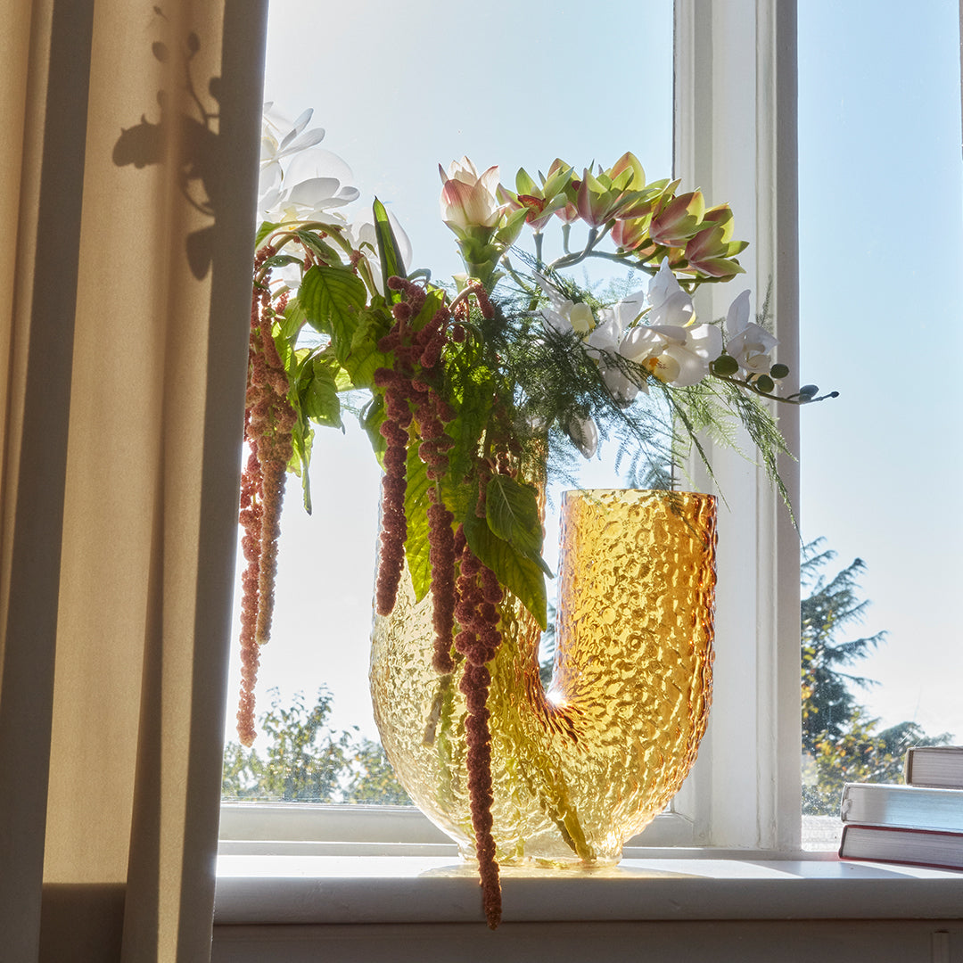 Clear glass vase in amber hue with a textured pattern, showcasing vibrant flowers and greenery against a bright window.