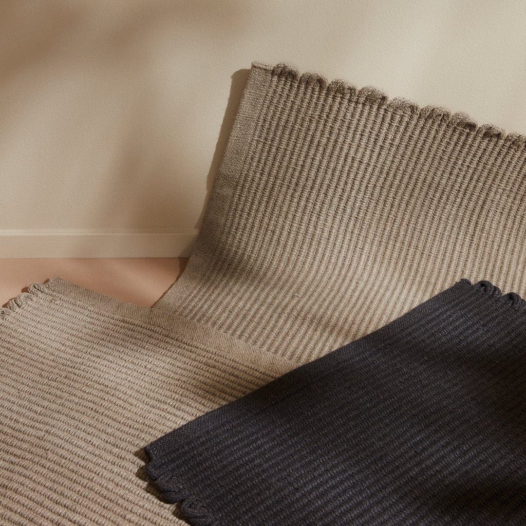 Stack of textured rugs in beige and navy blue with subtle ribbed patterns and fringed edges. Perfect for modern decor.