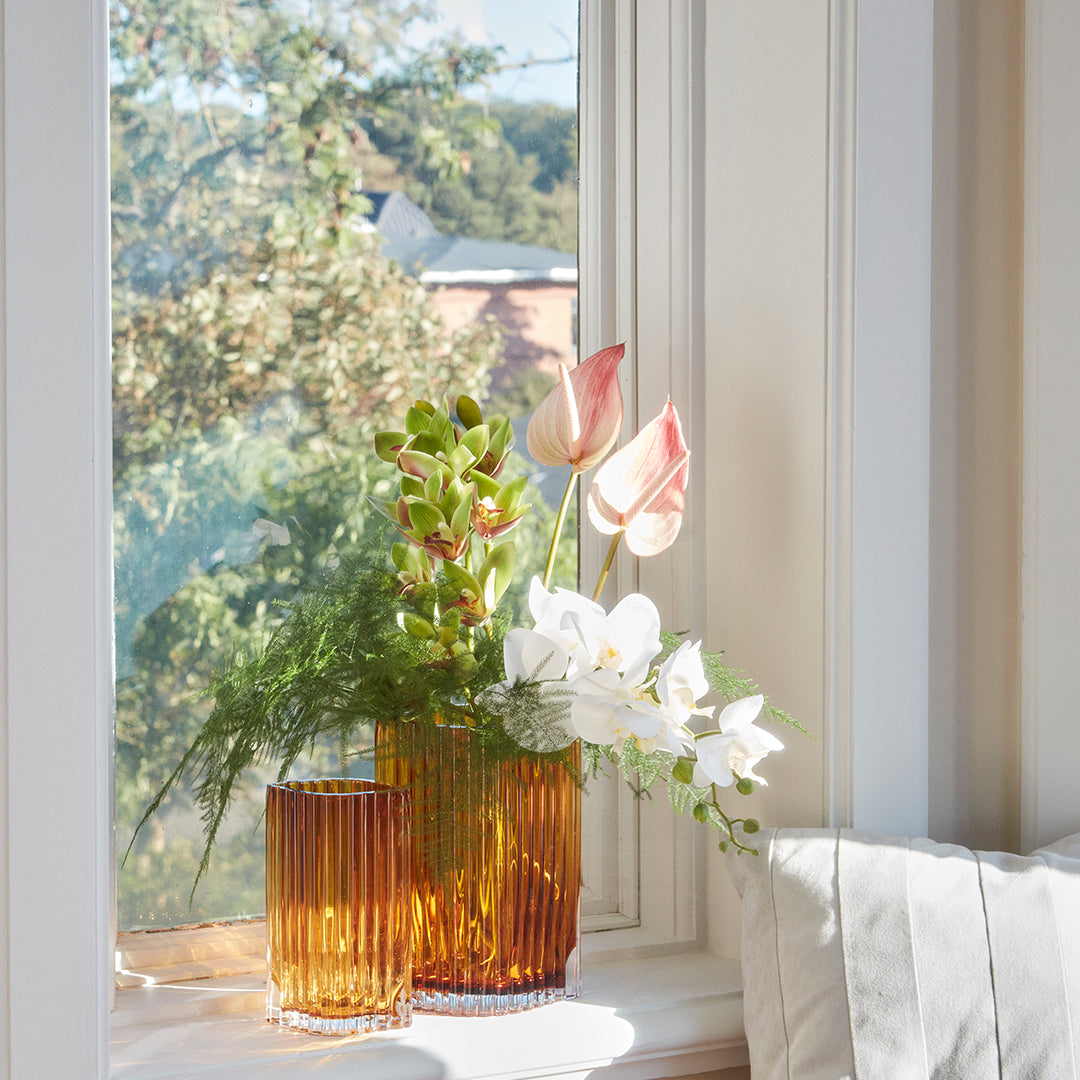 Amber ribbed glass vases with pink and white flowers in a bright window setting. Green foliage accentuates the colors.