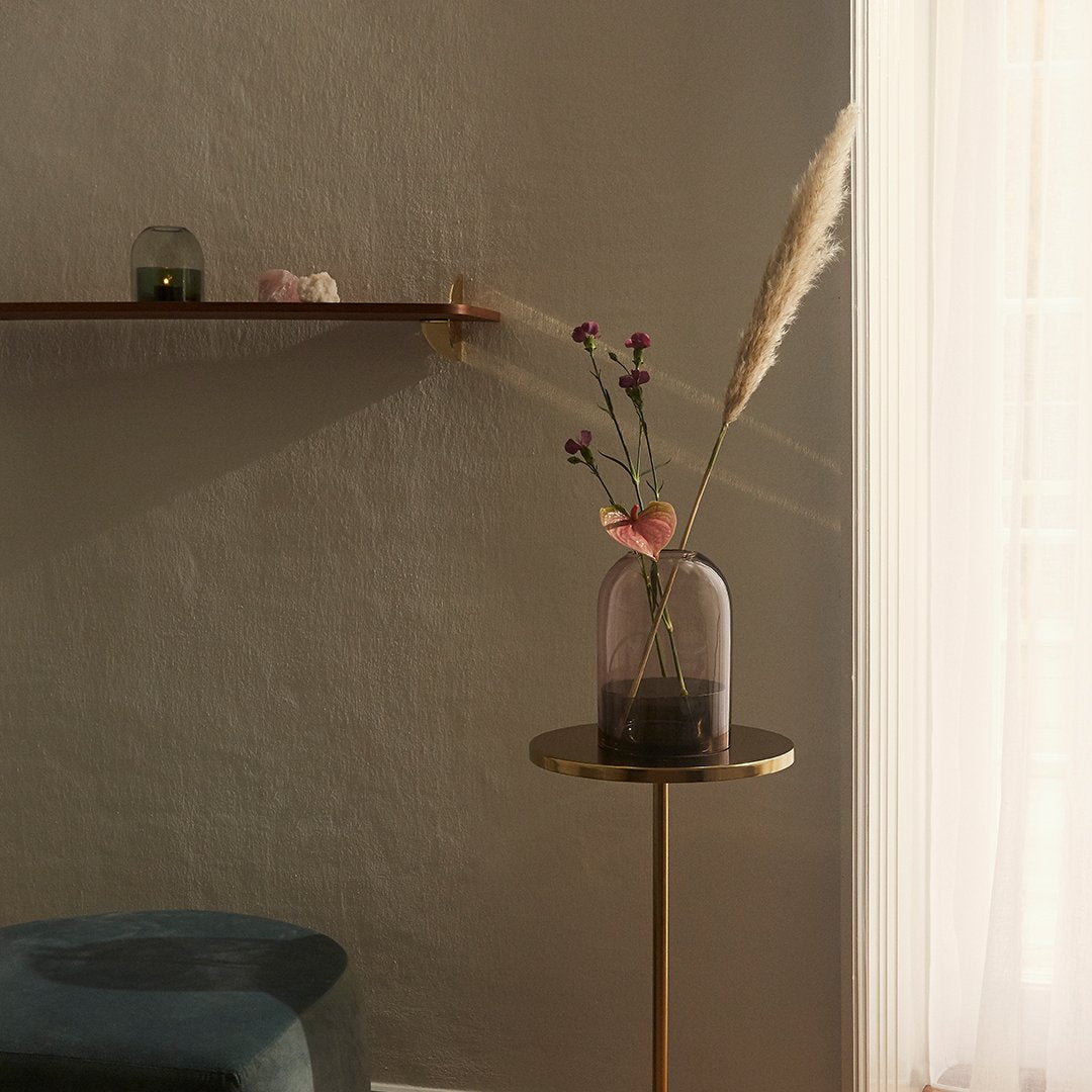 Pink transparent vase with golden accents, containing dried flowers, against a soft gray wall and light curtain backdrop.