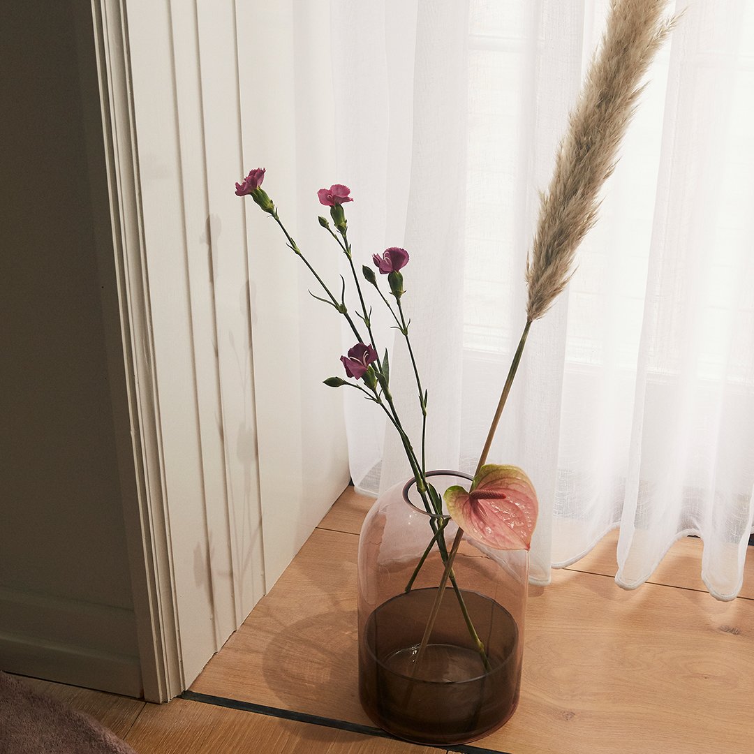 "Pink carnations and beige pampas grass in a translucent brown vase against a light backdrop."