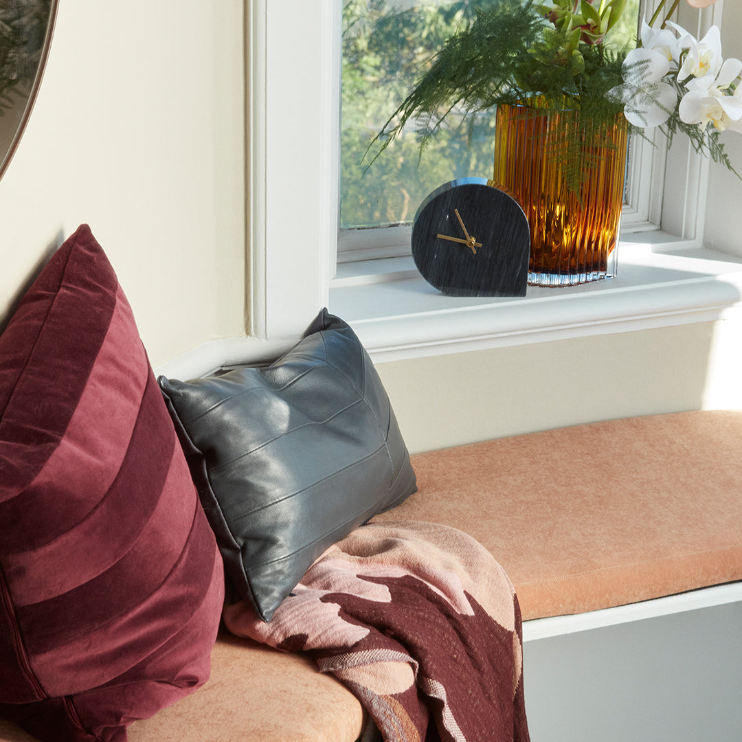 Burgundy and gray pillows on a soft pink bench, accented by a floral-patterned throw and a vibrant amber vase.