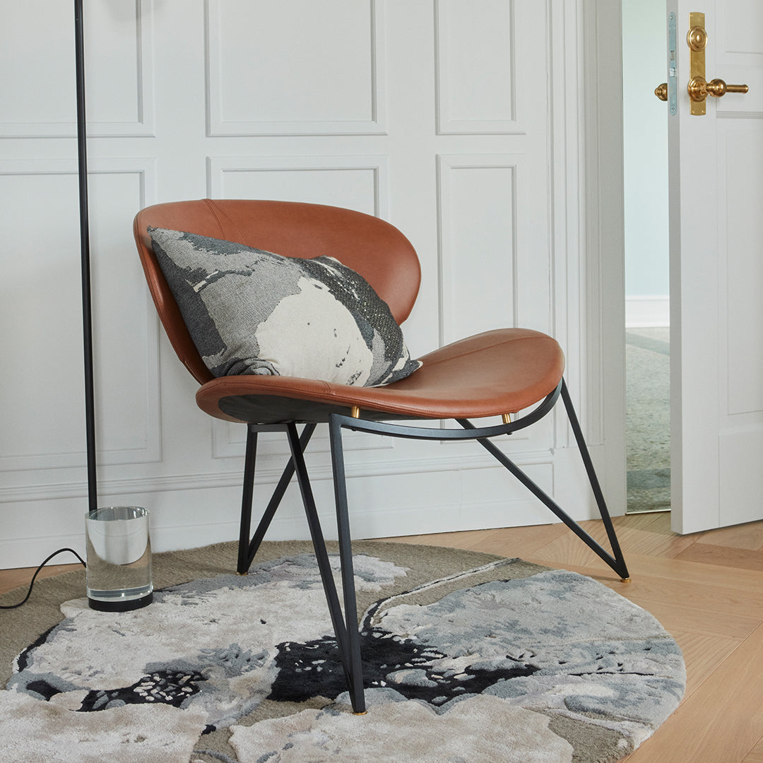Brown leather chair with geometric black metal legs, featuring a gray and white patterned pillow on a round abstract rug.