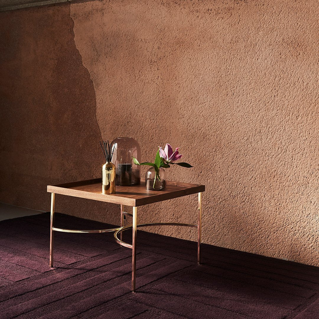 Wooden coffee table with gold legs, set against a warm, textured pink wall and deep burgundy rug. Elegant floral accents.