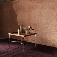 Wooden coffee table with gold legs, set against a warm, textured pink wall and deep burgundy rug. Elegant floral accents.