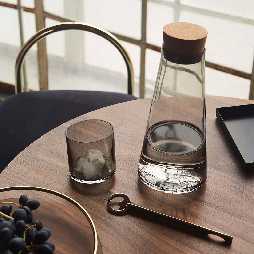 "Clear glass carafe with cork lid, dark gray glass tumbler, and metallic accents on a wooden table."