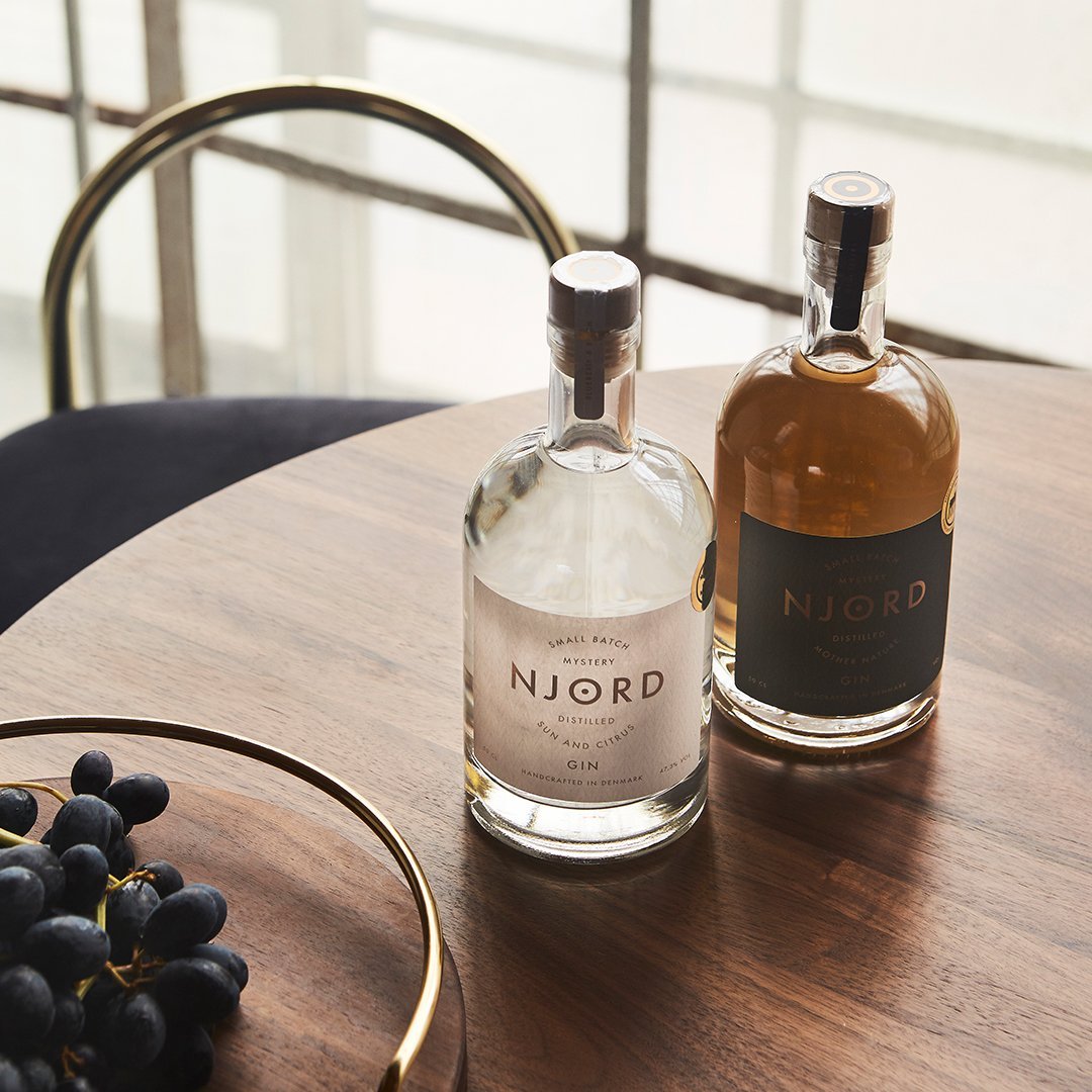 Two elegant glass bottles with clear and dark amber liquids, set on a wooden table with a gold circle and deep blue accents.