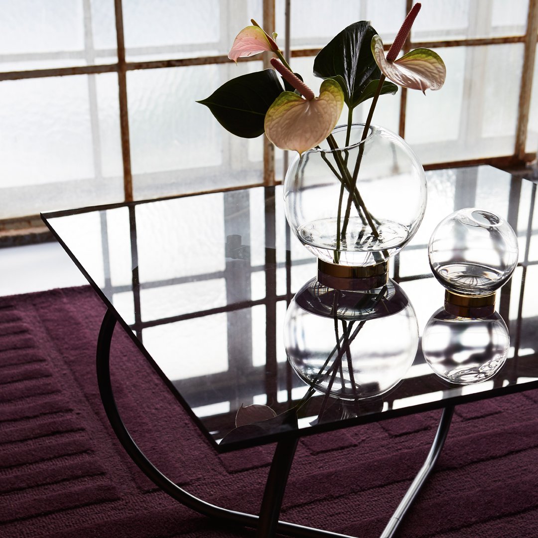 Black glass table atop a textured deep purple rug, featuring a clear glass vase with green and pink foliage.