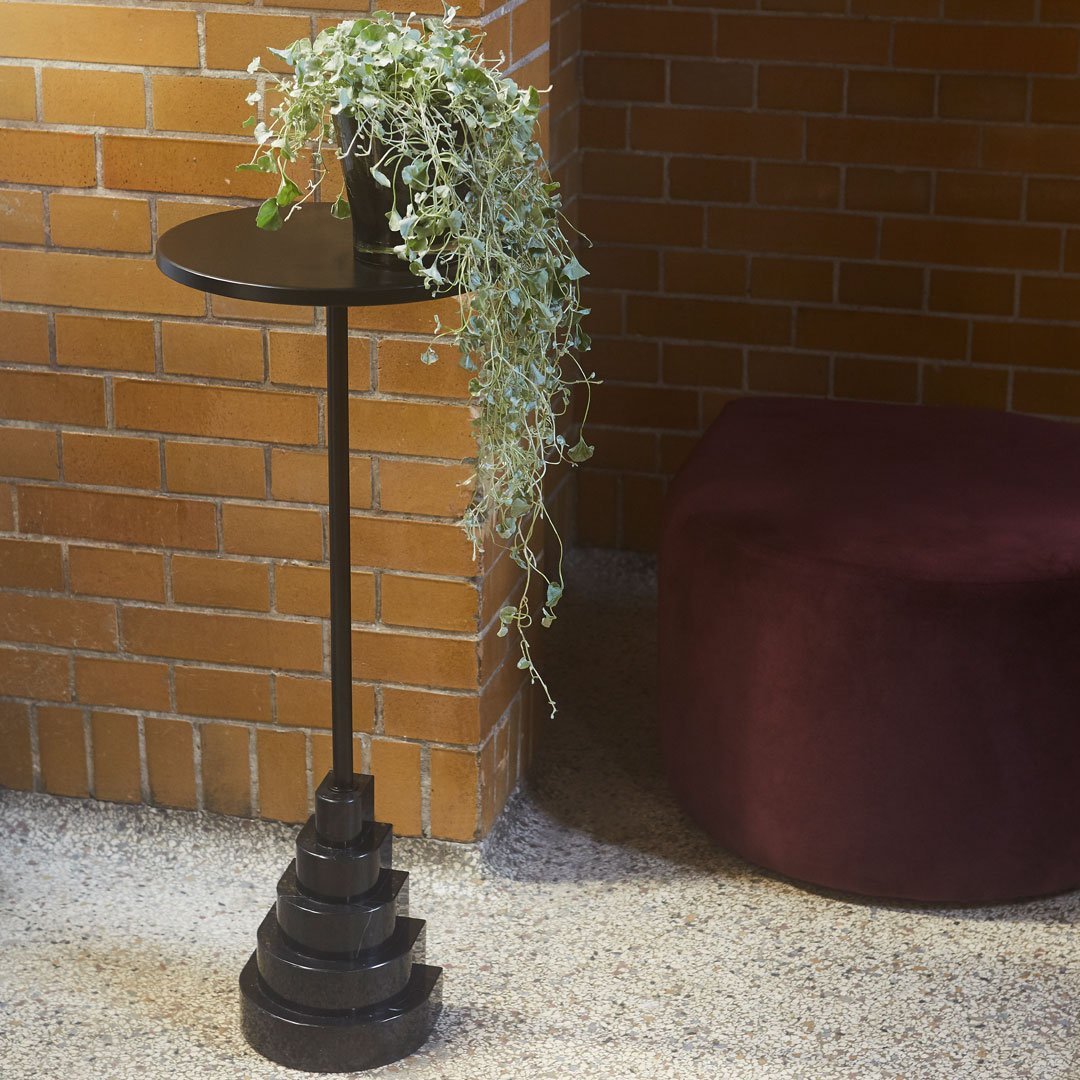 Black spiral plant stand with a circular top, accompanied by trailing greenery, set against a brick wall and plush purple ottoman.