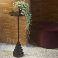 Black spiral plant stand with a circular top, accompanied by trailing greenery, set against a brick wall and plush purple ottoman.