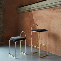 "Black upholstered stools with sleek gold metal frames against a warm, textured terracotta wall."