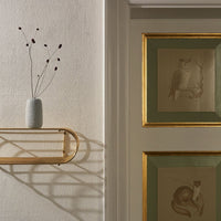 "Textured white vase with dark stems, against a soft beige wall and a gold-trimmed green frame with vintage art."