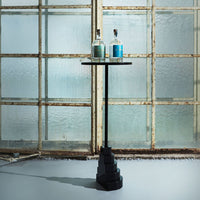 Stylish black pedestal table with a smooth round surface, set against a textured glass backdrop, featuring blue bottles.