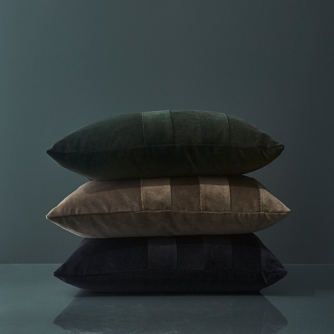 Stack of three plush pillows in dark green, earthy brown, and deep black with subtle panel patterns.