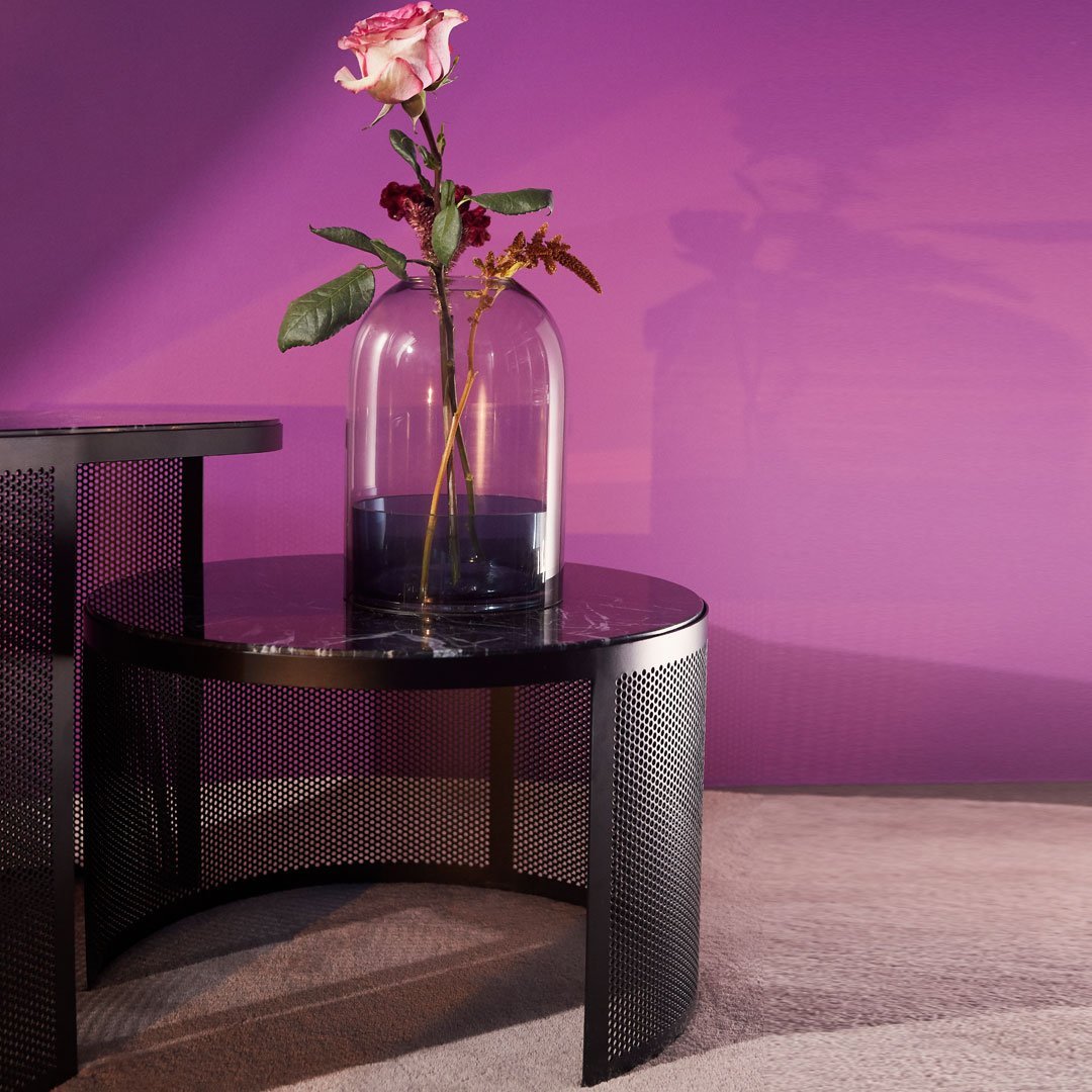 Black round table with textured pattern, featuring a clear vase and a pink rose against a vibrant purple wall.