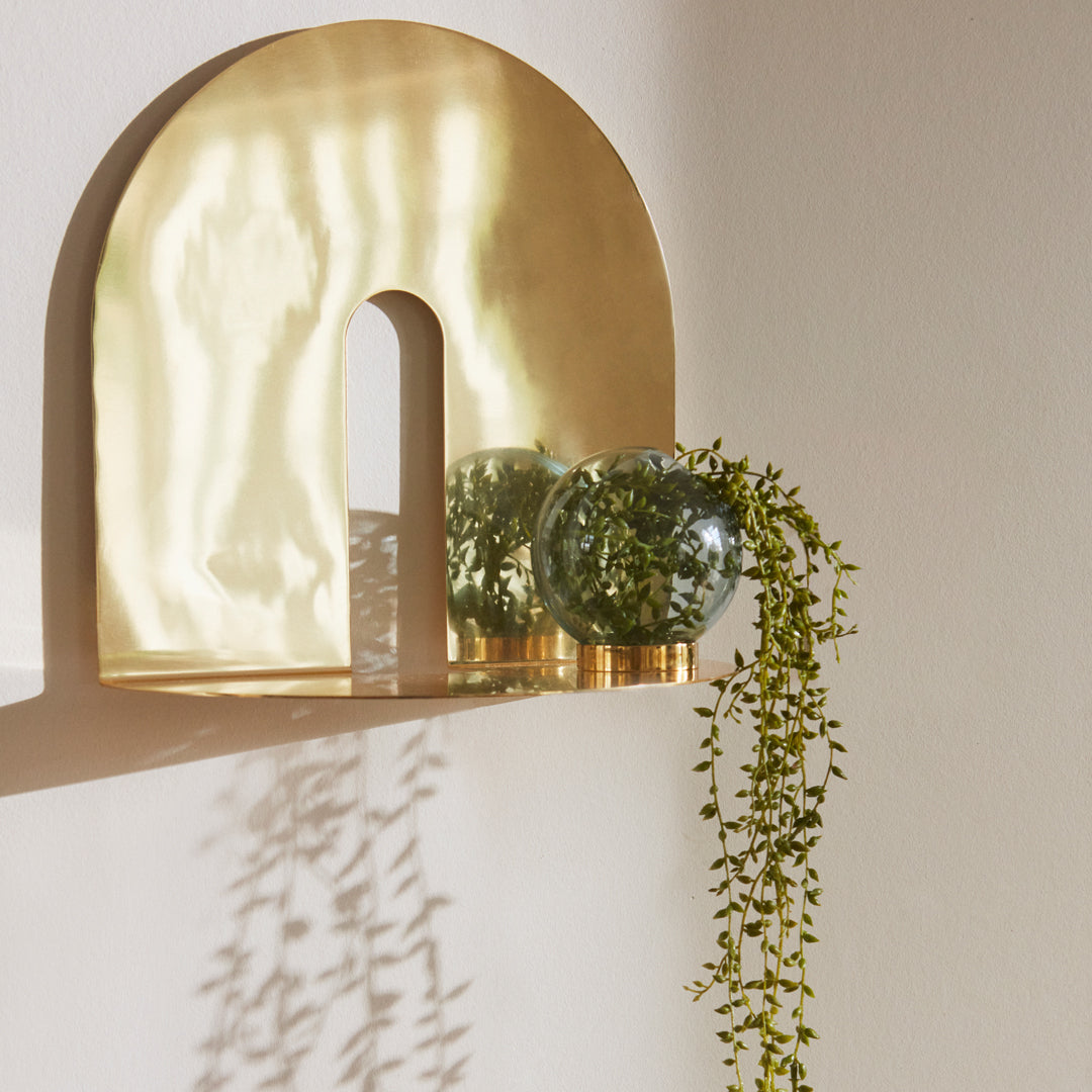 Brushed gold shelf with green glass vases and trailing vines casting soft shadows on a light background.