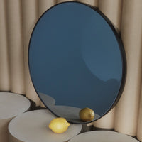 Round mirror with a sleek black frame, reflecting a soft blue background and a light yellow lemon on a neutral surface.