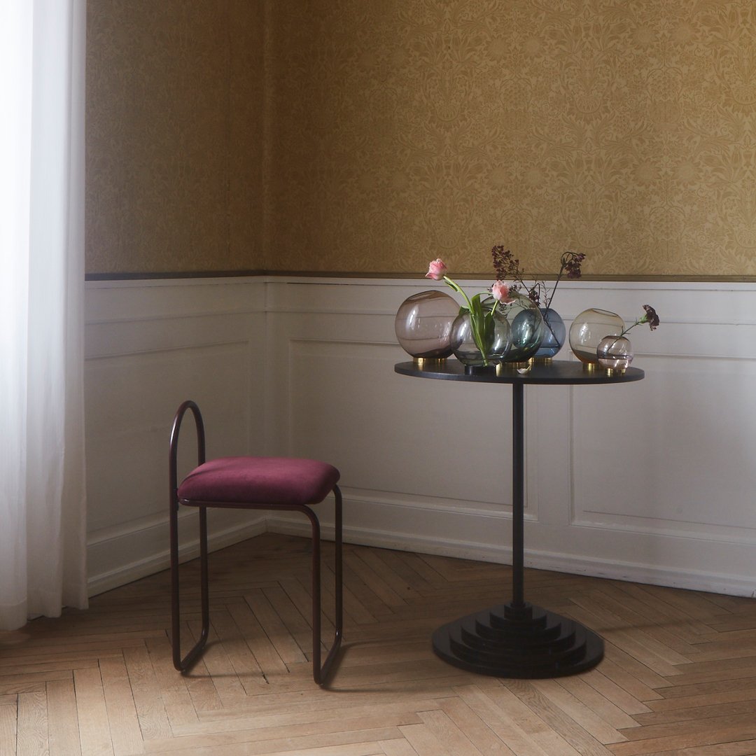 Modern black table with a round top, featuring a deep burgundy chair, set against a textured gold wallpaper backdrop.