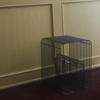 Black wireframe side table with sleek lines, set against a muted gold wall and deep red carpet.