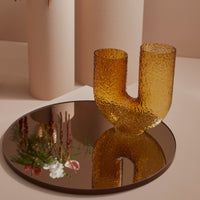 Amber textured glass vase with a unique U-shape, reflecting light on a round mirrored tray. Soft neutral background.