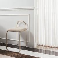 Beige upholstered stool with a sleek metal frame, set against a light wall and modern patterned flooring.