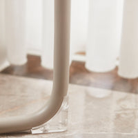 Beige curved leg with transparent base on a marble surface, complemented by soft white curtains in the background.