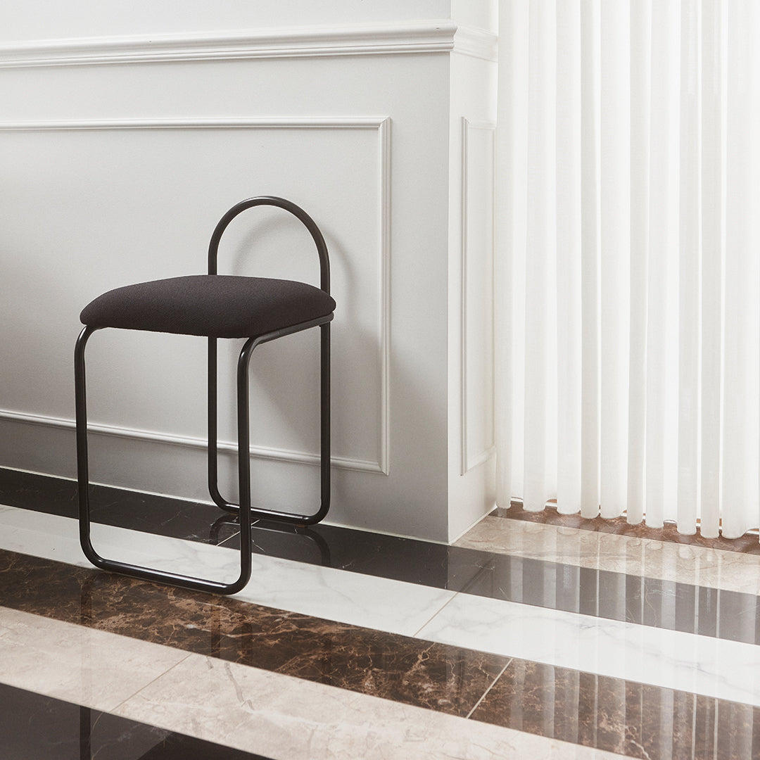 Black upholstered stool with a curved backrest, set against a black and white striped marble floor.