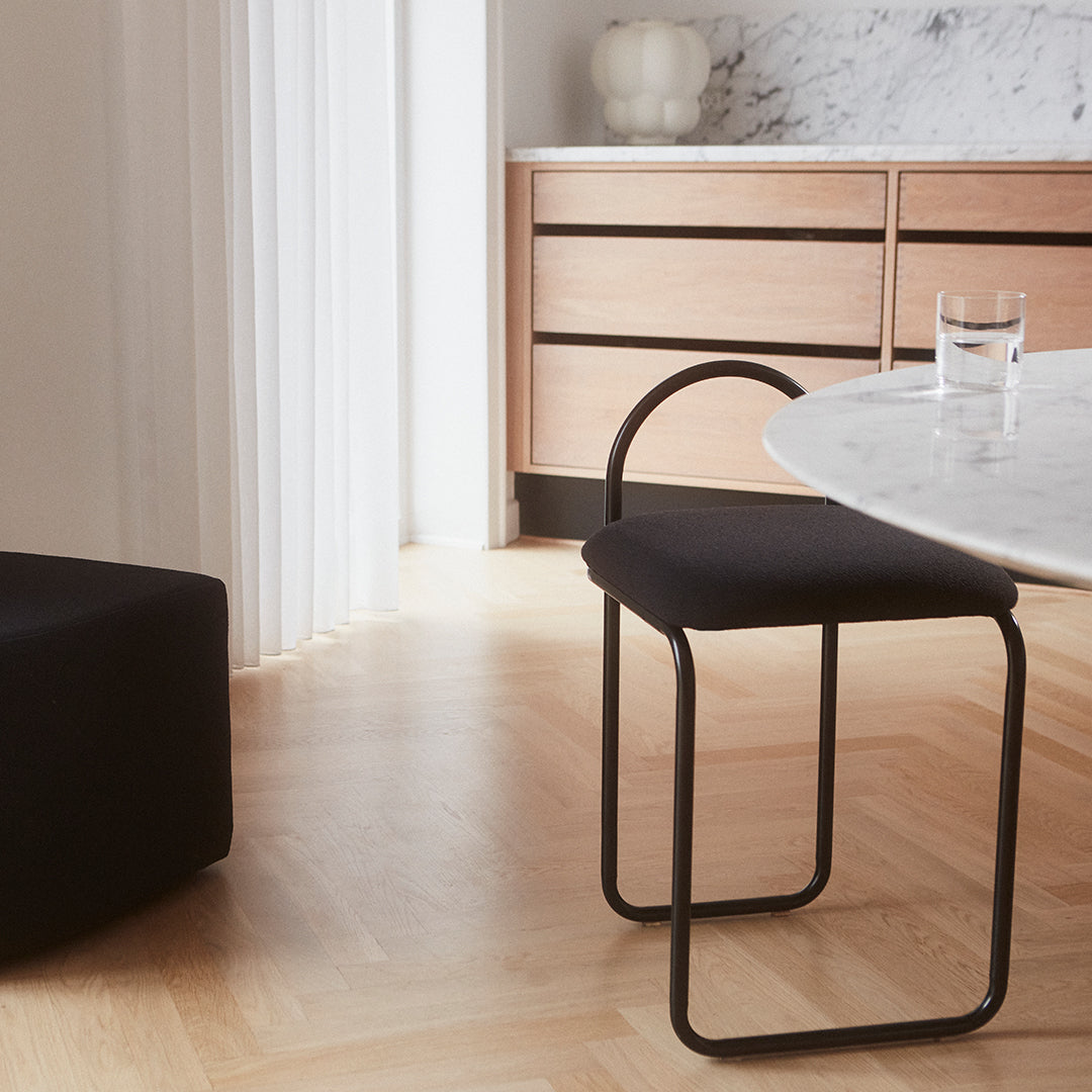 "Black minimalist chair with curved backrest, set on light wood floor beside marble table and wooden furniture."