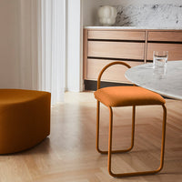 Orange upholstered chair with metal legs beside a mustard circular ottoman and a marble tabletop in a light, airy room.