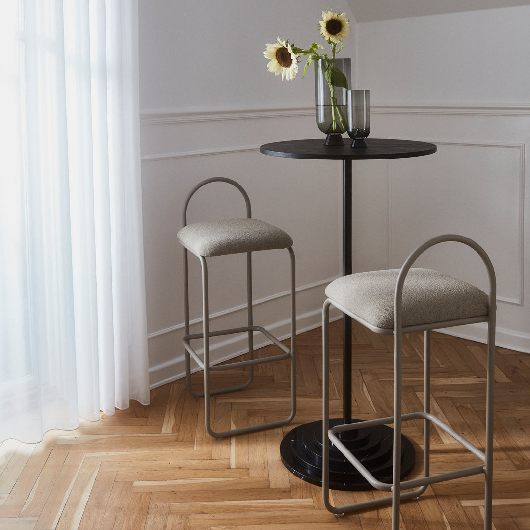 Elegant black round table with two light beige bar stools, set against a warm wooden floor and soft white curtains.