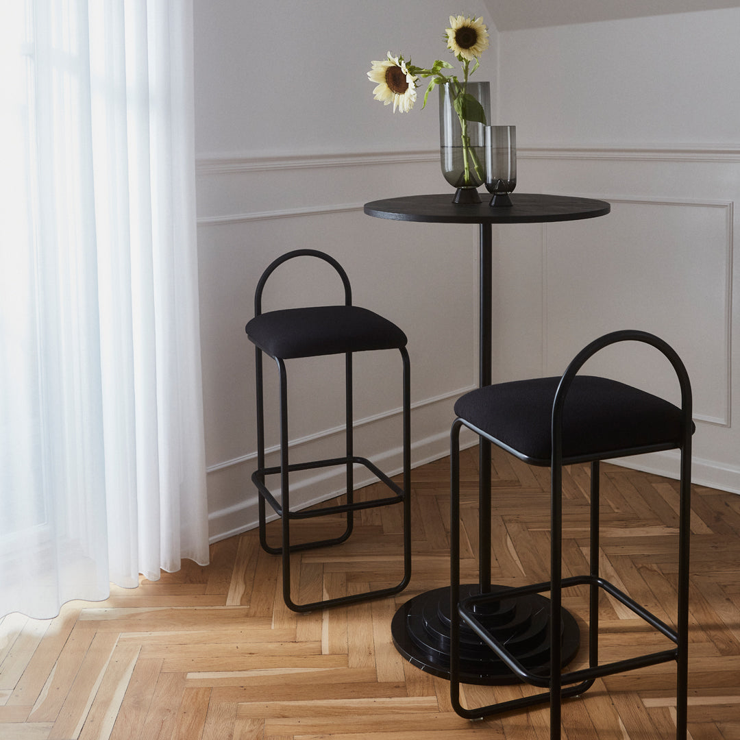 Black metal bar table and stools with a minimalist design, featuring a sleek round tabletop and soft, cushioned seat.