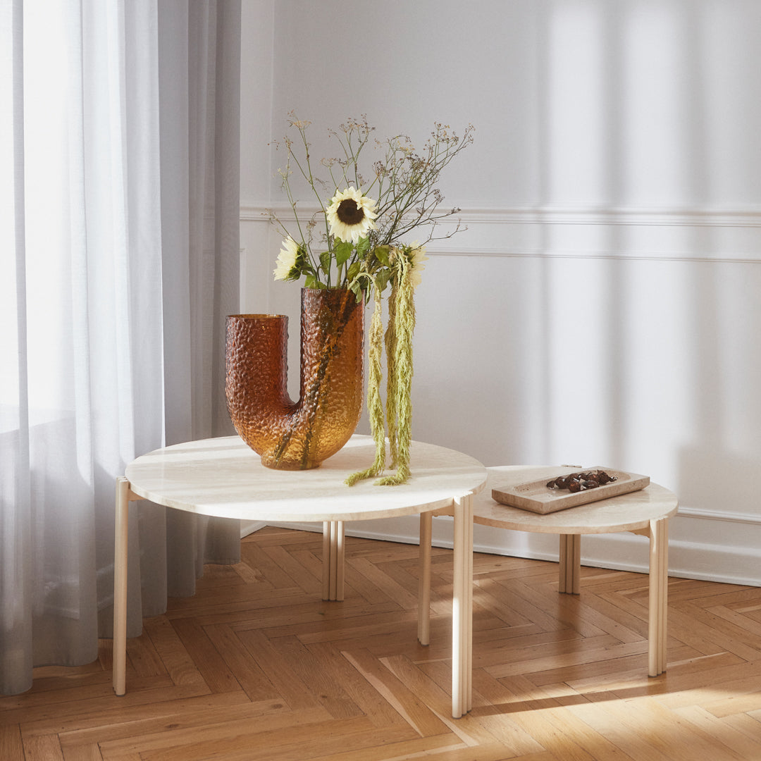 "Round light wood coffee table with a white top, accompanied by a textured amber vase and green floral arrangement."