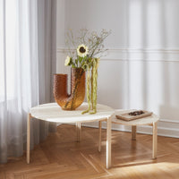 "Round light wood coffee table with a white top, accompanied by a textured amber vase and green floral arrangement."