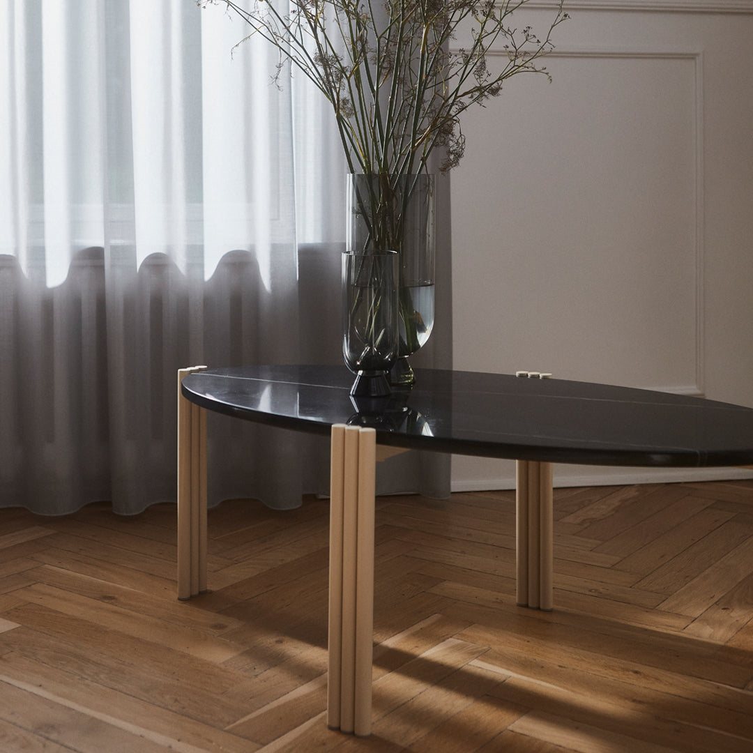 Black oval coffee table with light wood legs, set against a neutral-toned wooden floor and sheer gray curtains.