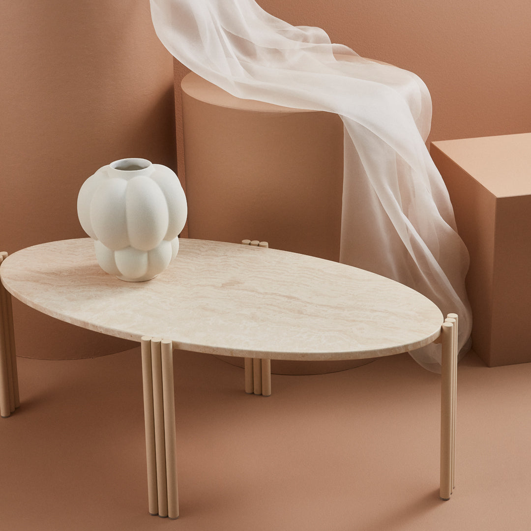 Elegant beige oval coffee table with light wood legs, accompanied by a soft white vase against a muted peach backdrop.